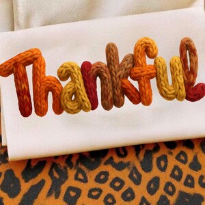 May include: A white rectangular decorative pillow with the word "THANKFUL" in 3D knitted letters. The letters are in shades of orange, yellow, and brown. The pillow is on a surface with a leopard print pattern.