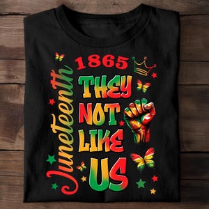 May include: Black t-shirt featuring a vibrant Juneteenth design. The graphic includes the text "1865 They Not Like Us", a raised fist, butterflies, stars, and a crown. The word "Juneteenth" is written vertically in green and red.