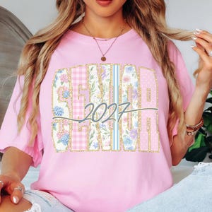 May include: Pink t-shirt with a large "SENIOR 2027" graphic. The text is in a gold outline, with floral, polka dot, and plaid patterns. The shirt is a light pink color.