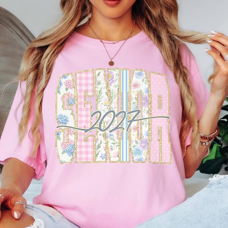 May include: Pink t-shirt with a large "SENIOR 2027" graphic. The text is in a gold outline, with floral, polka dot, and plaid patterns. The shirt is a light pink color.