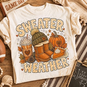 May include: A cream-colored t-shirt featuring a graphic print with the text "SWEATER WEATHER" in a dotted font. The design includes a coffee cup, a knit beanie, a pair of boots, a pumpkin, and a slice of pumpkin pie surrounded by autumn leaves.