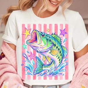 Op de afbeelding: Wit T-shirt met een kleurrijke illustratie van een vis met open mond, omringd door waterspetters en sterren. Het ontwerp is geplaatst tegen een achtergrond van roze en witte verticale strepen, wat een levendige en opvallende afbeelding cre&euml;ert.
