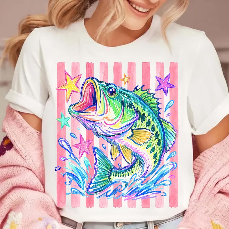 Op de afbeelding: Wit T-shirt met een kleurrijke illustratie van een vis met open mond, omringd door waterspetters en sterren. Het ontwerp is geplaatst tegen een achtergrond van roze en witte verticale strepen, wat een levendige en opvallende afbeelding cre&euml;ert.