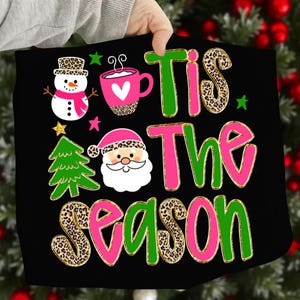 May include: Black Christmas-themed graphic with the phrase "Tis The Season" in green and pink. The design includes a snowman, a mug with a heart, a Christmas tree, and Santa Claus, all with leopard print accents.