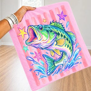 Op de afbeelding: Roze deken met een kleurrijke illustratie van een largemouth bass. De vis is afgebeeld met levendige groene, blauwe, gele en roze tinten, omringd door sterren en waterspetters. De achtergrond heeft verticale roze strepen.