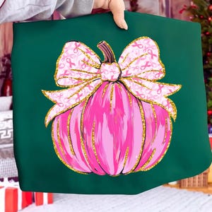 May include: A dark green tote bag featuring a pink pumpkin design with a bow. The bow is decorated with pink ribbons, and the pumpkin has gold glitter accents. The design is centered on the tote bag.