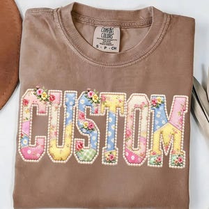 May include: A brown Comfort Colors t-shirt with the word "CUSTOM" appliquéd across the front. The letters are patchwork style, with floral and patterned fabrics, and are outlined with small pearls. Small floral accents are on top of each letter.
