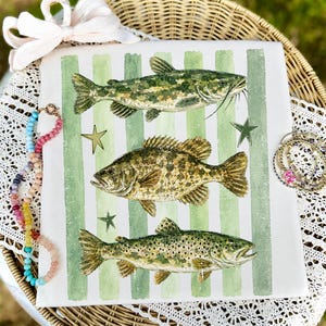May include: A white square decorative pillow featuring three detailed illustrations of fish: a catfish, a bass, and a trout. The design includes green vertical stripes and gold star accents. The pillow is placed on a wicker surface.