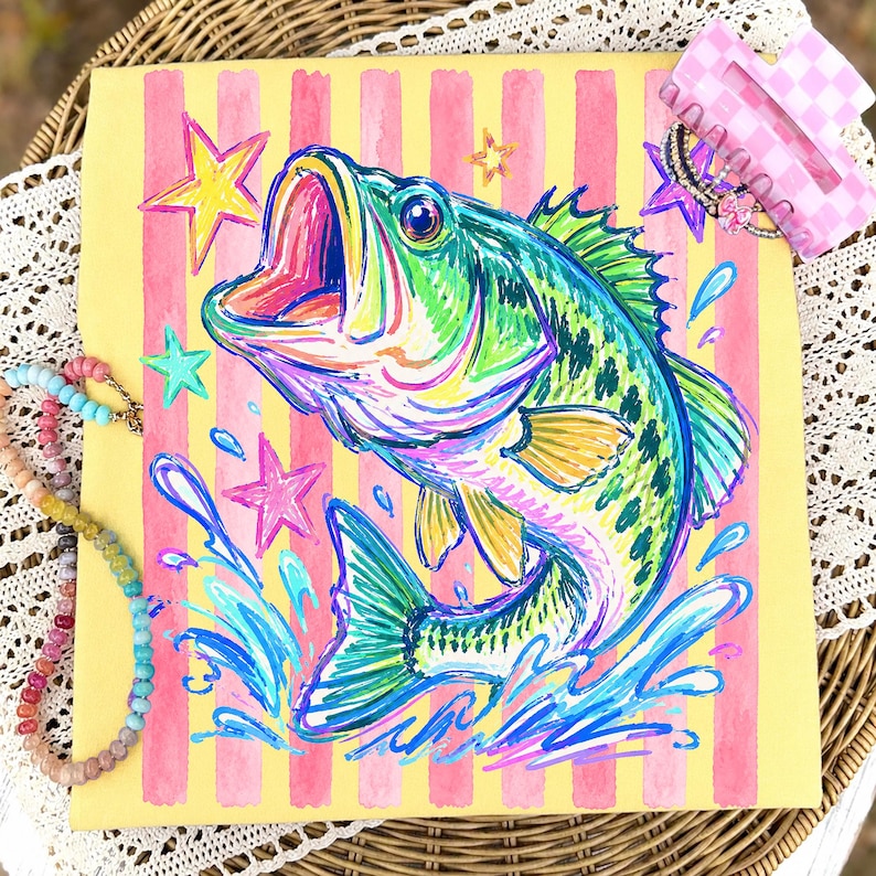 Op de afbeelding: Kleurrijke viskunst op een gele achtergrond met roze strepen en sterren. De vis is afgebeeld in levendige groen-, blauw- en geeltinten, met waterspetters. Een roze en witte haarclip en een kralenketting zijn ook zichtbaar.