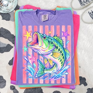 Op de afbeelding: Een stapel kleurrijke t-shirts. Het bovenste shirt is paars met een afbeelding van een kleurrijke vis met open mond, omringd door sterren en waterspetters. Het shirt heeft roze en witte verticale strepen. De andere shirts zijn roze, groen en oranje.