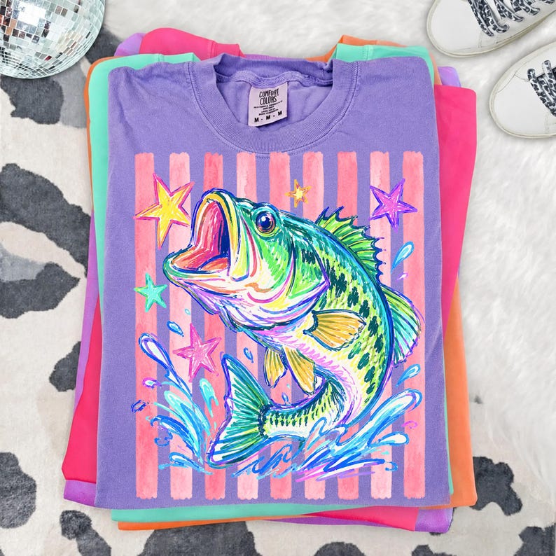 Op de afbeelding: Een stapel kleurrijke t-shirts. Het bovenste shirt is paars met een afbeelding van een kleurrijke vis met open mond, omringd door sterren en waterspetters. Het shirt heeft roze en witte verticale strepen. De andere shirts zijn roze, groen en oranje.