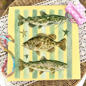 May include: A square, yellow canvas with green vertical stripes features three detailed illustrations of fish. The fish are a catfish, a bass, and a trout. Gold star accents are scattered around the fish. A pink and white checkered hair clip and a beaded necklace are in the background.