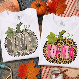 Pode incluir: Duas camisetas brancas com estampas gráficas de abóboras com padrões de leopardo. A abóbora da esquerda tem o nome OLIVER em letras bege e a da direita tem o nome SOPHIA em letras cor-de-rosa. Ambas as camisas possuem etiquetas Comfort Colors.