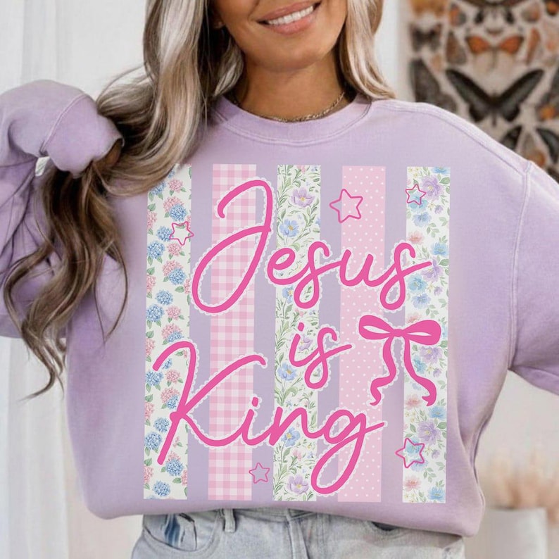 K&ouml;nnte beinhalten: Ein helllila Sweatshirt mit dem rosa Schriftzug "Jesus is King". Das Design zeigt vertikale Streifen aus floralen Mustern, Vichy-Karo und Tupfen. Kleine Sterne sind um den Text herum verstreut.
