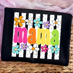 May include: Black t-shirt with a white striped design and colorful flowers. The word "mama" is displayed in large, retro-style letters in pink, orange, yellow, and green. The shirt has a playful and cheerful design.