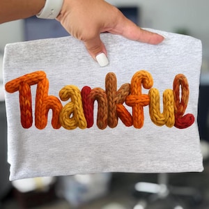 May include: A light gray t-shirt with the word "Thankful" in a 3D knitted style. The letters are in shades of orange, yellow, and brown. The shirt is held up by a hand with a white watch and white nail polish.