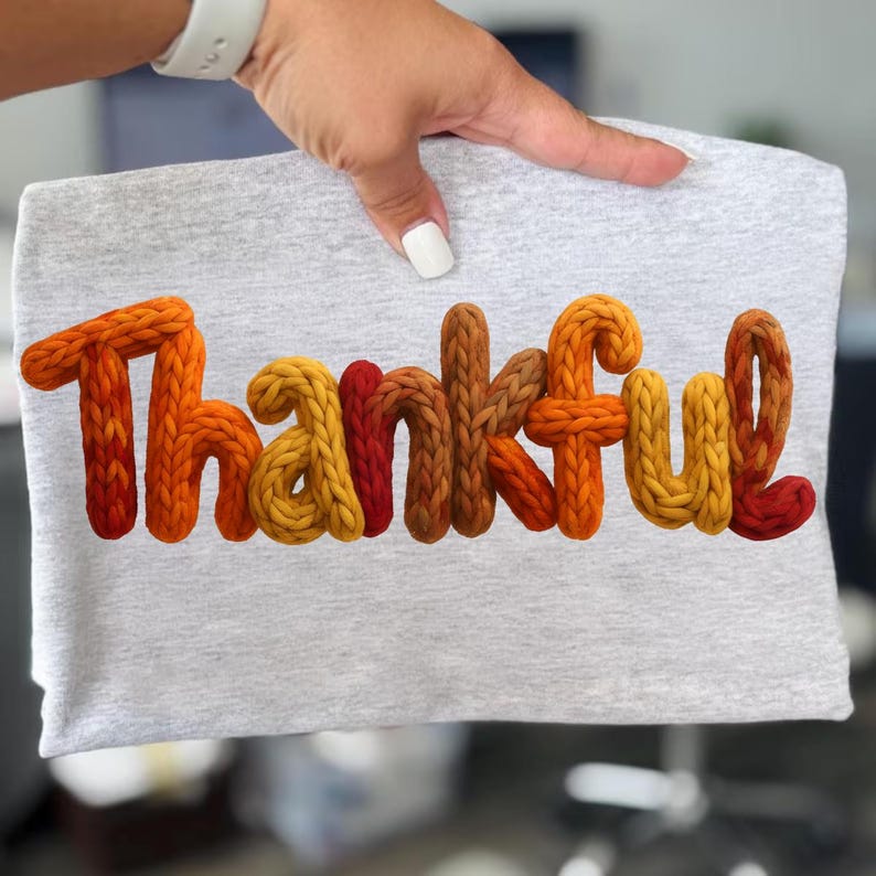 May include: A light gray t-shirt with the word "Thankful" in a 3D knitted style. The letters are in shades of orange, yellow, and brown. The shirt is held up by a hand with a white watch and white nail polish.