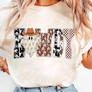 K&ouml;nnte beinhalten: Cremefarbenes T-Shirt mit dem Wort "Howdy" in einem Western-Design. Die Buchstaben sind mit Mustern wie Kuhdruck, Kakteen und einem Schachbrettmuster gef&uuml;llt. Ein Geist mit Cowboyhut befindet sich in der Mitte.