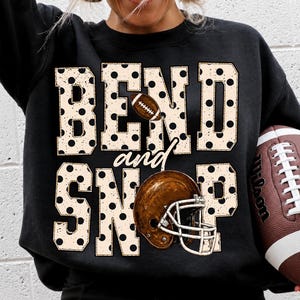 Puede incluir: Una sudadera negra de manga larga con el texto BEND and SNAP en grandes letras beige de bloque con lunares. El diseño incluye un gráfico de un balón de fútbol americano marrón y un casco de fútbol marrón con máscara blanca integrados.