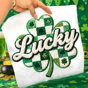 Op de afbeelding: Wit T-shirt met een groot, getextureerd klaverbladontwerp en het woord "Lucky" in een donzige, cursieve letter. Het klaverblad is groen en wit, met een geblokt patroon en kleine klaverbladaccenten, perfect voor St. Patrick's Day.