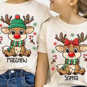 May include: Two cream-colored t-shirts with matching Christmas reindeer designs. One reindeer wears a green hat and scarf, the other a red bow. The shirts have the names "Matthew" and "Sophia" printed below the reindeer.