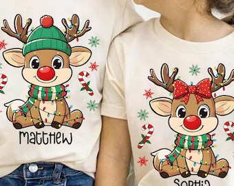 May include: Two cream-coloured t-shirts featuring Christmas reindeer designs. One reindeer has a green hat and scarf, the other a red bow. The shirts have the names "Matthew" and "Sophia" printed below the reindeer.