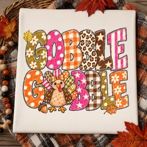 May include: A white fabric square featuring the text GOBBLE GOBBLE in large, multi-patterned letters. The patterns include daisies, pink polka dots, orange gingham, leopard print, brown stars, and stripes. A cartoon turkey is centered in the text.