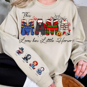 Pu&ograve; includere: Una felpa beige con la scritta "This Mama Loves her Little Horror". La parola "Mama" &egrave; disegnata con personaggi di film horror. La manica presenta personaggi horror dei cartoni animati. Un capo d'abbigliamento a tema Halloween.