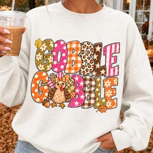 May include: A white crewneck sweatshirt featuring the text GOBBLE GOBBLE in large, multi-patterned letters. The design includes floral, polka dot, plaid, leopard, and star prints in orange, pink, brown, and yellow, with a cartoon turkey graphic.