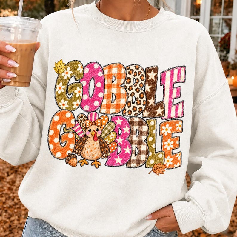 May include: A white crewneck sweatshirt featuring the text GOBBLE GOBBLE in large, multi-patterned letters. The design includes floral, polka dot, plaid, leopard, and star prints in orange, pink, brown, and yellow, with a cartoon turkey graphic.