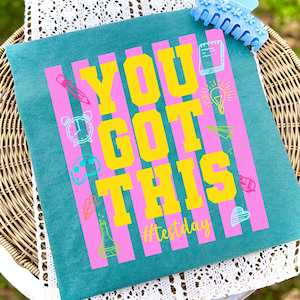 Peut inclure: Coussin carré turquoise avec le texte "YOU GOT THIS" en lettres jaunes, sur des rayures verticales roses. La phrase "#testday" est en dessous. Le motif comprend de petites illustrations d'objets scolaires.
