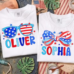 Puede incluir: Dos camisetas blancas con diseños patrióticos. Una camiseta presenta gafas de sol azules con un diseño de estrellas y rayas y el nombre "OLIVER" en rojo y azul. La otra camiseta tiene un diseño de pajarita con estrellas y rayas y el nombre "SOPHIA".