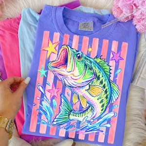 Op de afbeelding: Een paars Comfort Colors T-shirt met een kleurrijke illustratie van een vis, sterren en waterspetters. Het ontwerp is geplaatst op roze verticale strepen. Andere pastelkleurige shirts zijn ernaast opgevouwen.