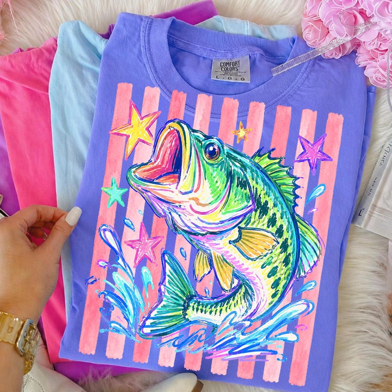 Op de afbeelding: Een paars Comfort Colors T-shirt met een kleurrijke illustratie van een vis, sterren en waterspetters. Het ontwerp is geplaatst op roze verticale strepen. Andere pastelkleurige shirts zijn ernaast opgevouwen.