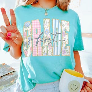 May include: A turquoise t-shirt with the words "GRADE FIRST" in a collegiate style. The letters are filled with floral and plaid patterns. The word "first" is in a cursive font. A yellow and white mug with a smiley face is in the image.