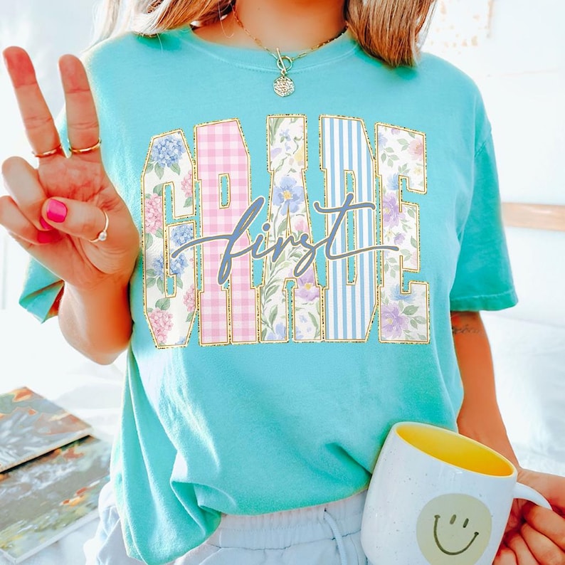 May include: A turquoise t-shirt with the words "GRADE FIRST" in a collegiate style. The letters are filled with floral and plaid patterns. The word "first" is in a cursive font. A yellow and white mug with a smiley face is in the image.