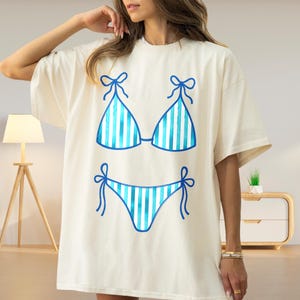 May include: Cream-coloured oversized t-shirt featuring a blue and white striped bikini design. The bikini top and bottom are outlined in blue with bow details. A casual and summery graphic tee.