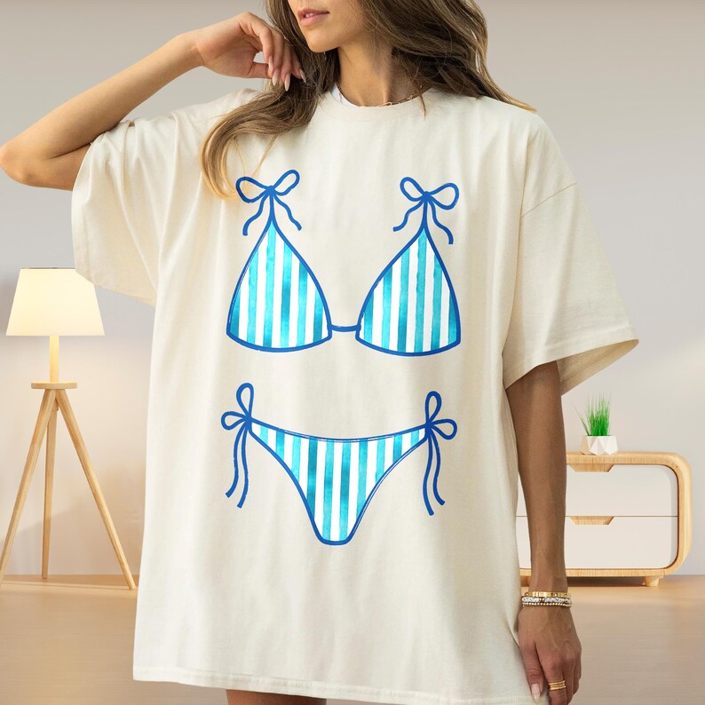 May include: Cream-coloured oversized t-shirt featuring a blue and white striped bikini design. The bikini top and bottom are outlined in blue with bow details. A casual and summery graphic tee.