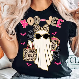 May include: Black t-shirt with a Halloween-themed graphic. The design features a ghost wearing sunglasses and a bow, holding a purse and a mug. The text "BOO-VEE" is above the ghost. Pink bats and spiderwebs complete the design.