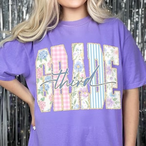May include: A lilac t-shirt featuring the word "GRADE" in large, decorative letters. The letters are filled with floral and gingham patterns, and the word "Third" is written in cursive across the centre.