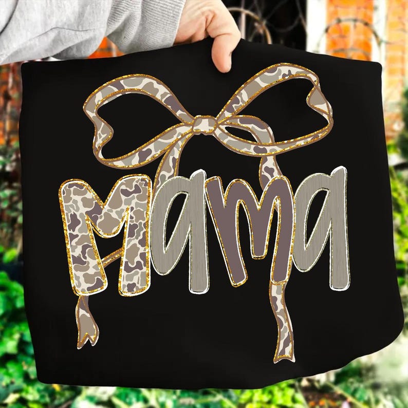 May include: Black tote bag with the word "MAMA" in a gray, outlined font. A bow with a camouflage pattern and gold trim is above the word. The bag is held by a person.