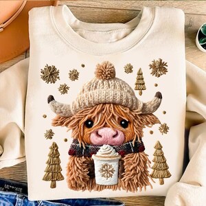 May include: Cream-colored sweatshirt featuring a Highland cow design. The cow wears a knit hat, scarf, and holds a coffee cup. Decorative snowflakes and Christmas trees surround the cow. A cozy and festive design.