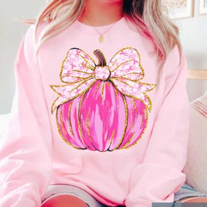 May include: Pink sweatshirt featuring a pink pumpkin design with a gold glitter outline and a bow adorned with pink ribbons. The pumpkin has a stem and is centered on the front of the shirt. The bow is decorated with pink ribbons.