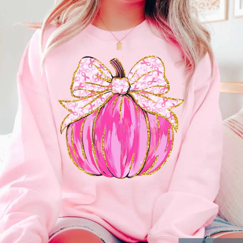 May include: Pink sweatshirt featuring a pink pumpkin design with a gold glitter outline and a bow adorned with pink ribbons. The pumpkin has a stem and is centered on the front of the shirt. The bow is decorated with pink ribbons.
