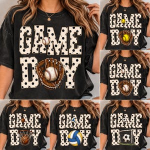 May include: A black short-sleeve t-shirt featuring the text GAME DAY in large white letters with black polka dots. The graphic includes various sports equipment illustrations such as baseballs, a baseball glove, a basketball, and a soccer ball.