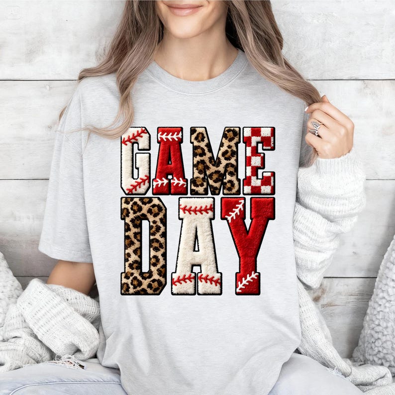 May include: A light gray sweatshirt with the words "GAME DAY" in a bold, multi-textured design. The letters feature baseball stitching, leopard print, and a red and white checkered pattern, creating a sporty and eye-catching look.