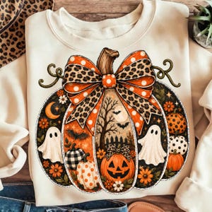 May include: A cream-coloured sweatshirt featuring a large graphic of a pumpkin decorated with a leopard print and orange polka dot bow. The pumpkin contains scenes of ghosts, bats, a moon, flowers, and smaller pumpkins with various patterns.
