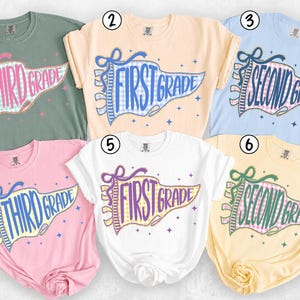 Könnte beinhalten: Sechs kurzärmelige T-Shirts mit Rundhalsausschnitt in verschiedenen Pastellfarben, jedes mit einer wimpelförmigen Grafik mit Schleife und Sternen. Die Designs zeigen den Text 'FIRST GRADE', 'SECOND GRADE' oder 'THIRD GRADE'.