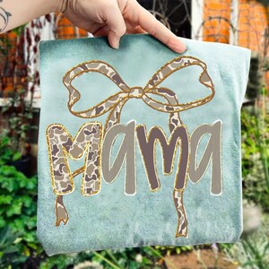 May include: A light blue decorative pillow with the word "MAMA" in a gray font with gold glitter accents. The letter "M" is filled with a camouflage pattern. A bow with a camouflage pattern and gold glitter accents is at the top.