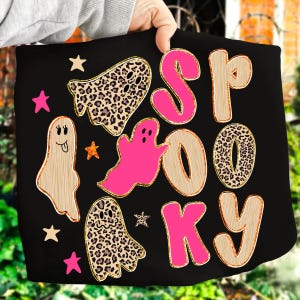 May include: Black tote bag with the word "SPOOKY" in pink and wood-grain letters. The design includes pink stars, ghosts, and leopard print ghosts. The bag is held by a person.